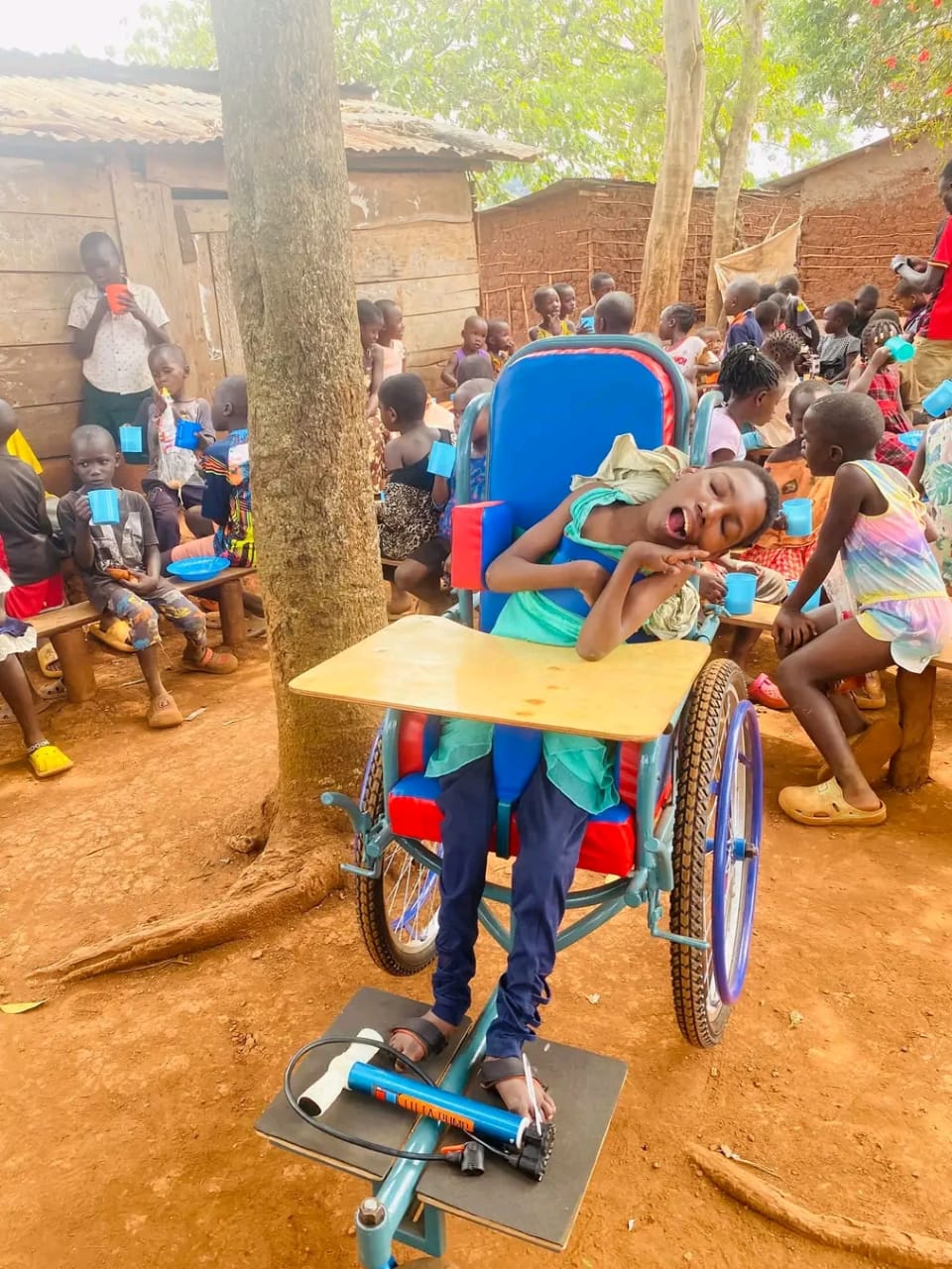 A child in a wheelchair at a DENO DEE FOUNDATION outreach, with many children sharing drinks behind